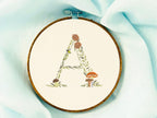 A letter A embroidered in a woodland theme with ferns, branches, acorn, a bee, and mushrooms in greens, browns, oranges, and yellow. This letter is stitched onto a white cotton background with a wooden embroidery hoop holding it. There are embroidery items like material, scissors, and thread around the photo. The photo is a top view.