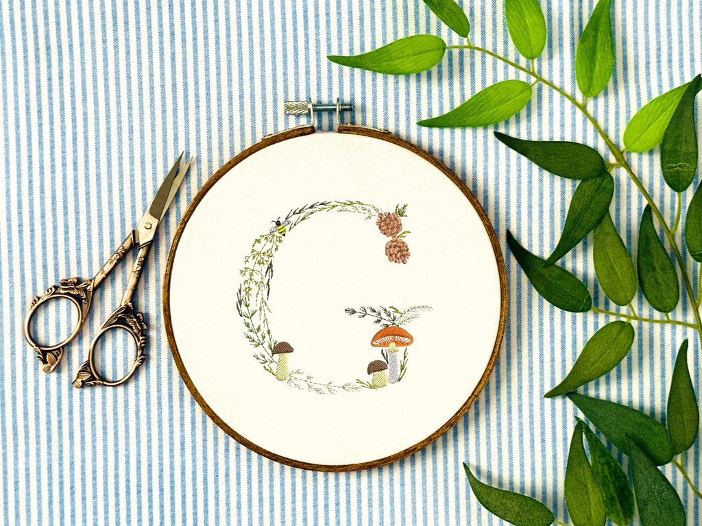 A letter G embroidered in a woodland theme with ferns, branches, acorn, a bee, and mushrooms in greens, browns, oranges, and yellow. This letter is stitched onto a white cotton background with a wooden embroidery hoop holding it. There are embroidery items like material, scissors, and thread around the photo. The photo is a top view.