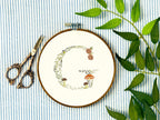 A letter G embroidered in a woodland theme with ferns, branches, acorn, a bee, and mushrooms in greens, browns, oranges, and yellow. This letter is stitched onto a white cotton background with a wooden embroidery hoop holding it. There are embroidery items like material, scissors, and thread around the photo. The photo is a top view.