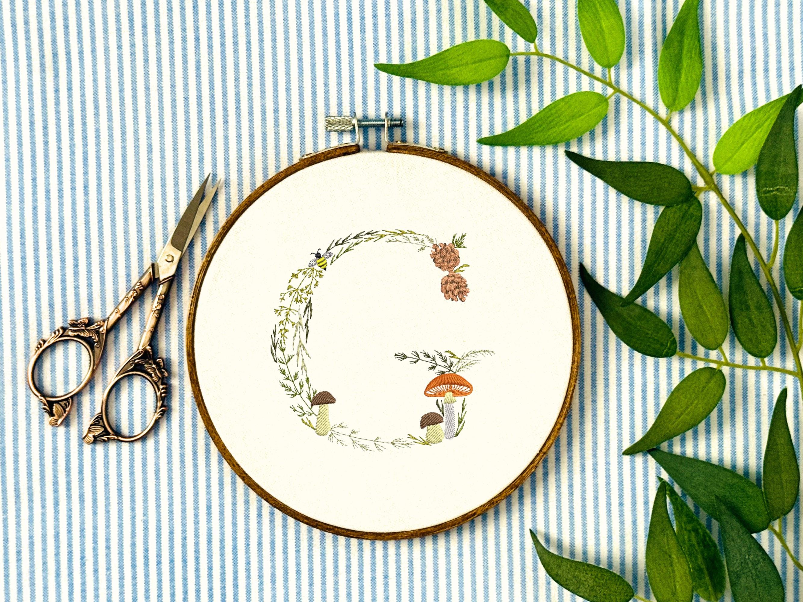 A letter G embroidered in a woodland theme with ferns, branches, acorn, a bee, and mushrooms in greens, browns, oranges, and yellow. This letter is stitched onto a white cotton background with a wooden embroidery hoop holding it. There are embroidery items like material, scissors, and thread around the photo. The photo is a top view.