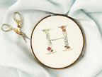 A letter H embroidered in a woodland theme with ferns, branches, acorn, a bee, and mushrooms in greens, browns, oranges, and yellow. This letter is stitched onto a white cotton background with a wooden embroidery hoop holding it.  The photo is a top view.