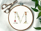 A letter M embroidered in a woodland theme with ferns, branches, acorn, a bee, and mushrooms in greens, browns, oranges, and yellow. This letter is stitched onto a white cotton background with a wooden embroidery hoop holding it. There are embroidery items like material, scissors, and thread around the photo. The photo is a top view.