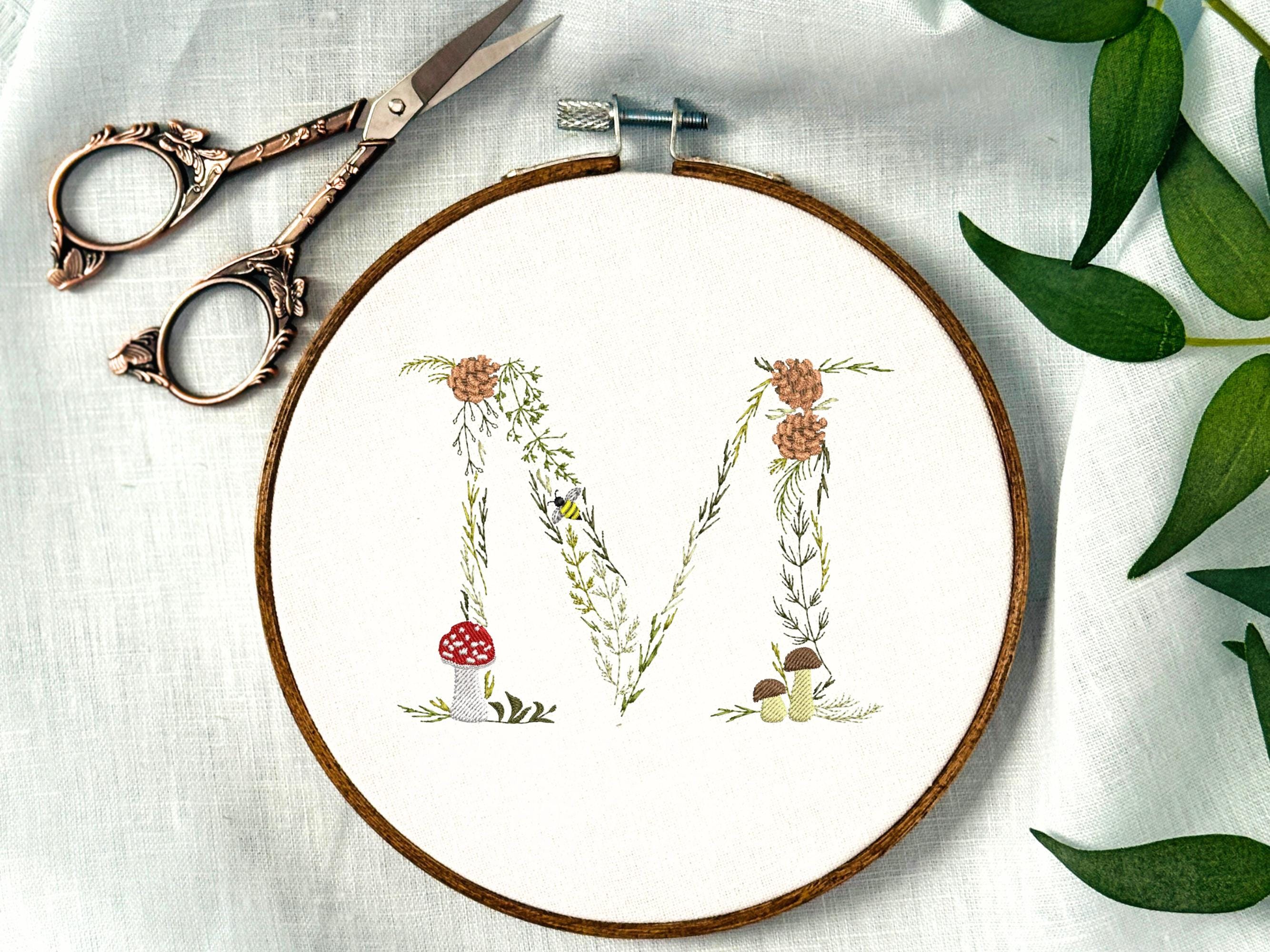 A letter M embroidered in a woodland theme with ferns, branches, acorn, a bee, and mushrooms in greens, browns, oranges, and yellow. This letter is stitched onto a white cotton background with a wooden embroidery hoop holding it. There are embroidery items like material, scissors, and thread around the photo. The photo is a top view.