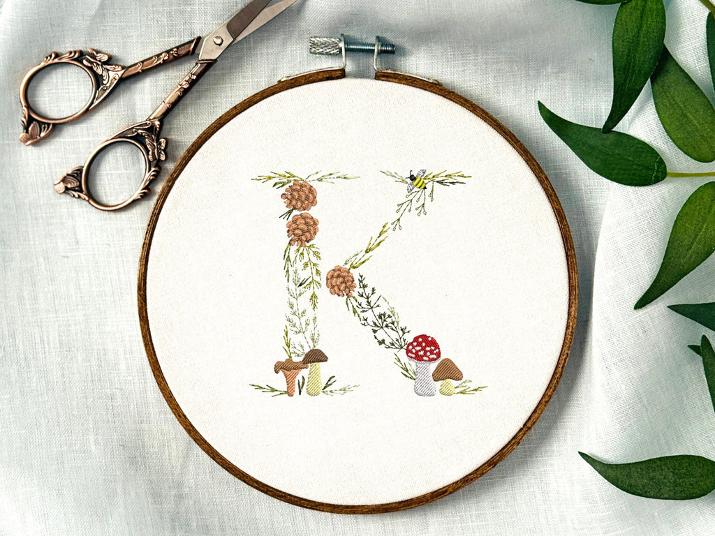 A letter K embroidered in a woodland theme with ferns, branches, acorn, a bee, and mushrooms in greens, browns, oranges, and yellow. This letter is stitched onto a white cotton background with a wooden embroidery hoop holding it. There are embroidery items like material, scissors, and thread around the photo. The photo is a top view.