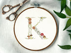 A letter K embroidered in a woodland theme with ferns, branches, acorn, a bee, and mushrooms in greens, browns, oranges, and yellow. This letter is stitched onto a white cotton background with a wooden embroidery hoop holding it. There are embroidery items like material, scissors, and thread around the photo. The photo is a top view.