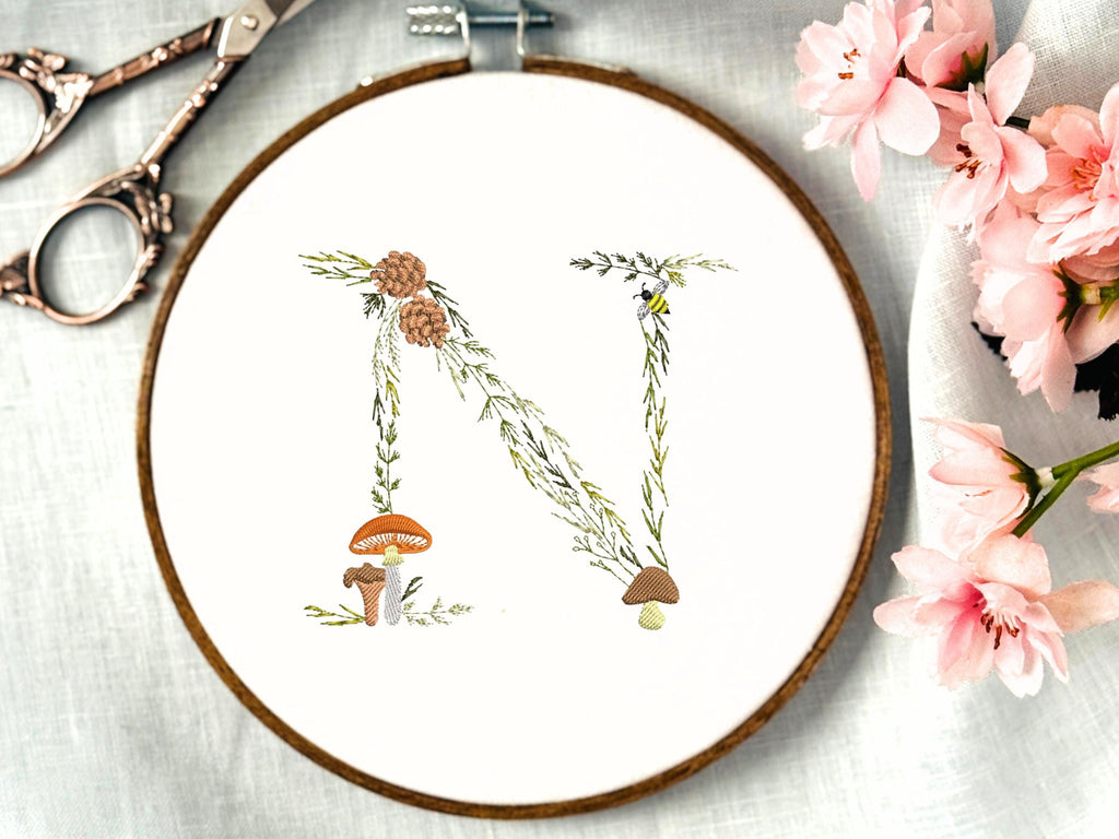 A letter N embroidered in a woodland theme with ferns, branches, acorn, a bee, and mushrooms in greens, browns, oranges, and yellow. This letter is stitched onto a white cotton background with a wooden embroidery hoop holding it. There are embroidery items like material, scissors, and thread around the photo. The photo is a top view.