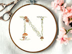 A letter N embroidered in a woodland theme with ferns, branches, acorn, a bee, and mushrooms in greens, browns, oranges, and yellow. This letter is stitched onto a white cotton background with a wooden embroidery hoop holding it. There are embroidery items like material, scissors, and thread around the photo. The photo is a top view.