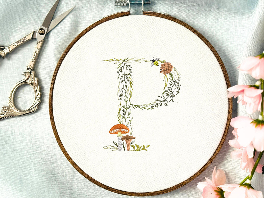 A letter P embroidered in a woodland theme with ferns, branches, acorn, a bee, and mushrooms in greens, browns, oranges, and yellow. This letter is stitched onto a white cotton background with a wooden embroidery hoop holding it. There are embroidery items like material, scissors, and thread around the photo. The photo is a top view.