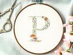 A letter P embroidered in a woodland theme with ferns, branches, acorn, a bee, and mushrooms in greens, browns, oranges, and yellow. This letter is stitched onto a white cotton background with a wooden embroidery hoop holding it. There are embroidery items like material, scissors, and thread around the photo. The photo is a top view.