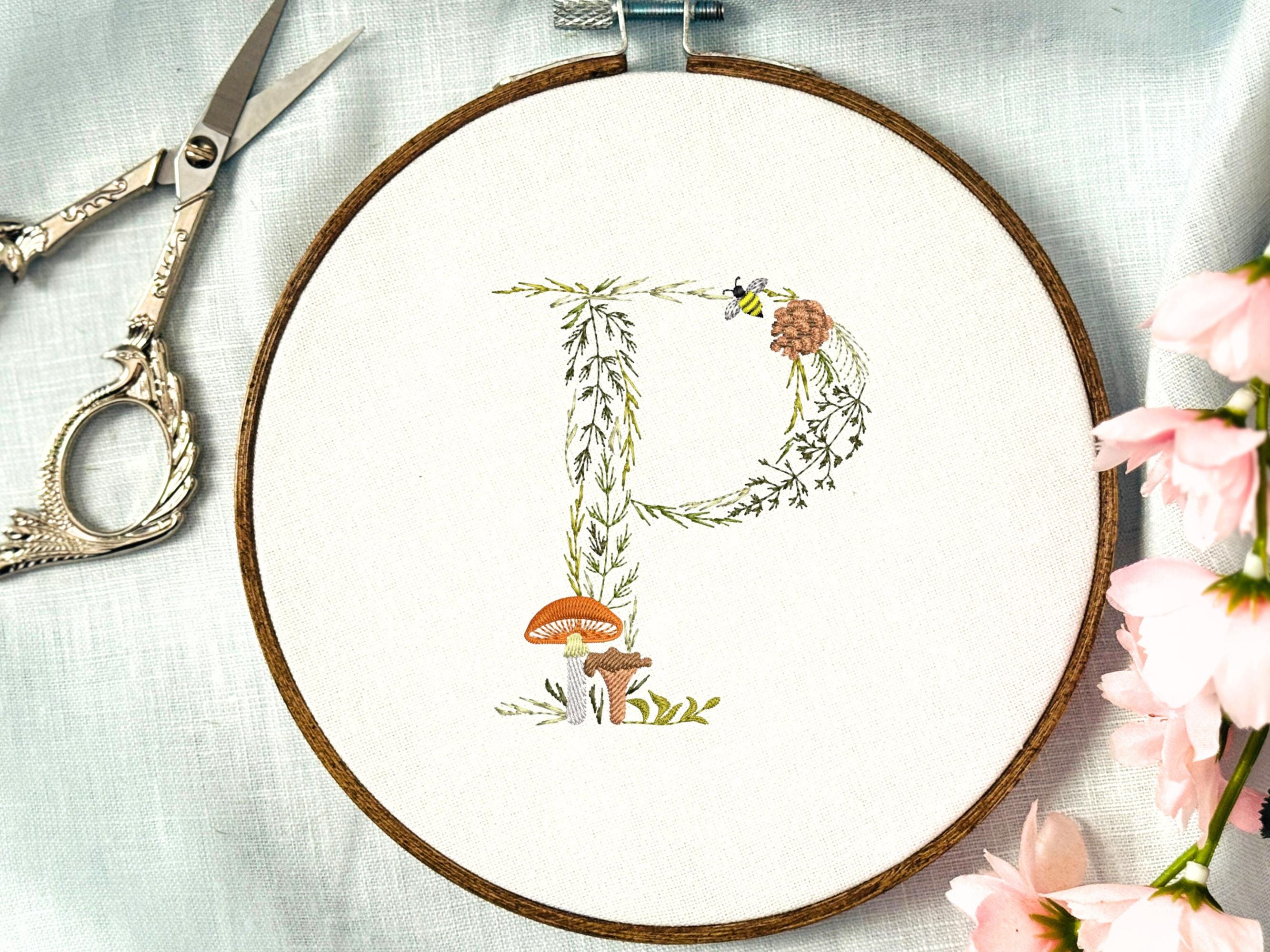 A letter P embroidered in a woodland theme with ferns, branches, acorn, a bee, and mushrooms in greens, browns, oranges, and yellow. This letter is stitched onto a white cotton background with a wooden embroidery hoop holding it. There are embroidery items like material, scissors, and thread around the photo. The photo is a top view.