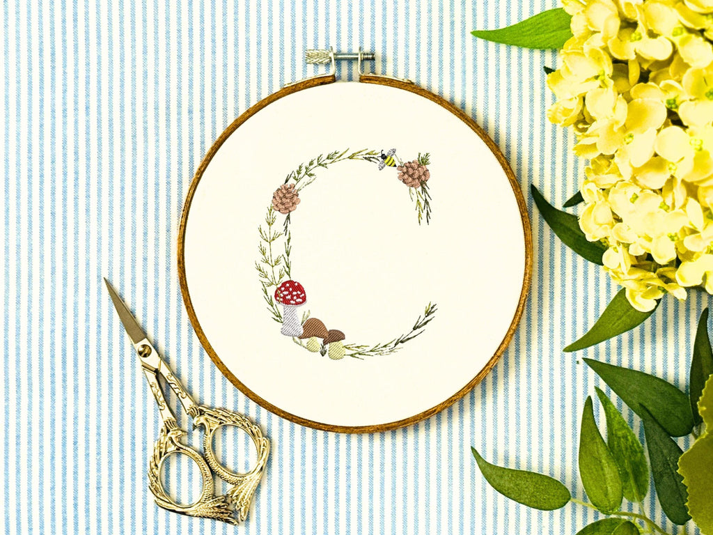 A letter C embroidered in a woodland theme with ferns, branches, acorn, a bee, and mushrooms in greens, browns, oranges, and yellow. This letter is stitched onto a white cotton background with a wooden embroidery hoop holding it. There are embroidery items like material, scissors, and thread around the photo. The photo is a top view.