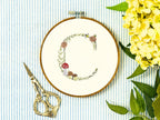 A letter C embroidered in a woodland theme with ferns, branches, acorn, a bee, and mushrooms in greens, browns, oranges, and yellow. This letter is stitched onto a white cotton background with a wooden embroidery hoop holding it. There are embroidery items like material, scissors, and thread around the photo. The photo is a top view.
