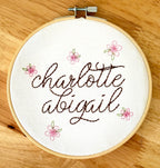 Light wood embroidery hoop art set on a wood table with the name Charlotte Abigail embroidered in dark brown. The hoop has five pink embroidered flowers around the name: Charlotte Abigail in cursive. It is stitched on white cotton fabric.