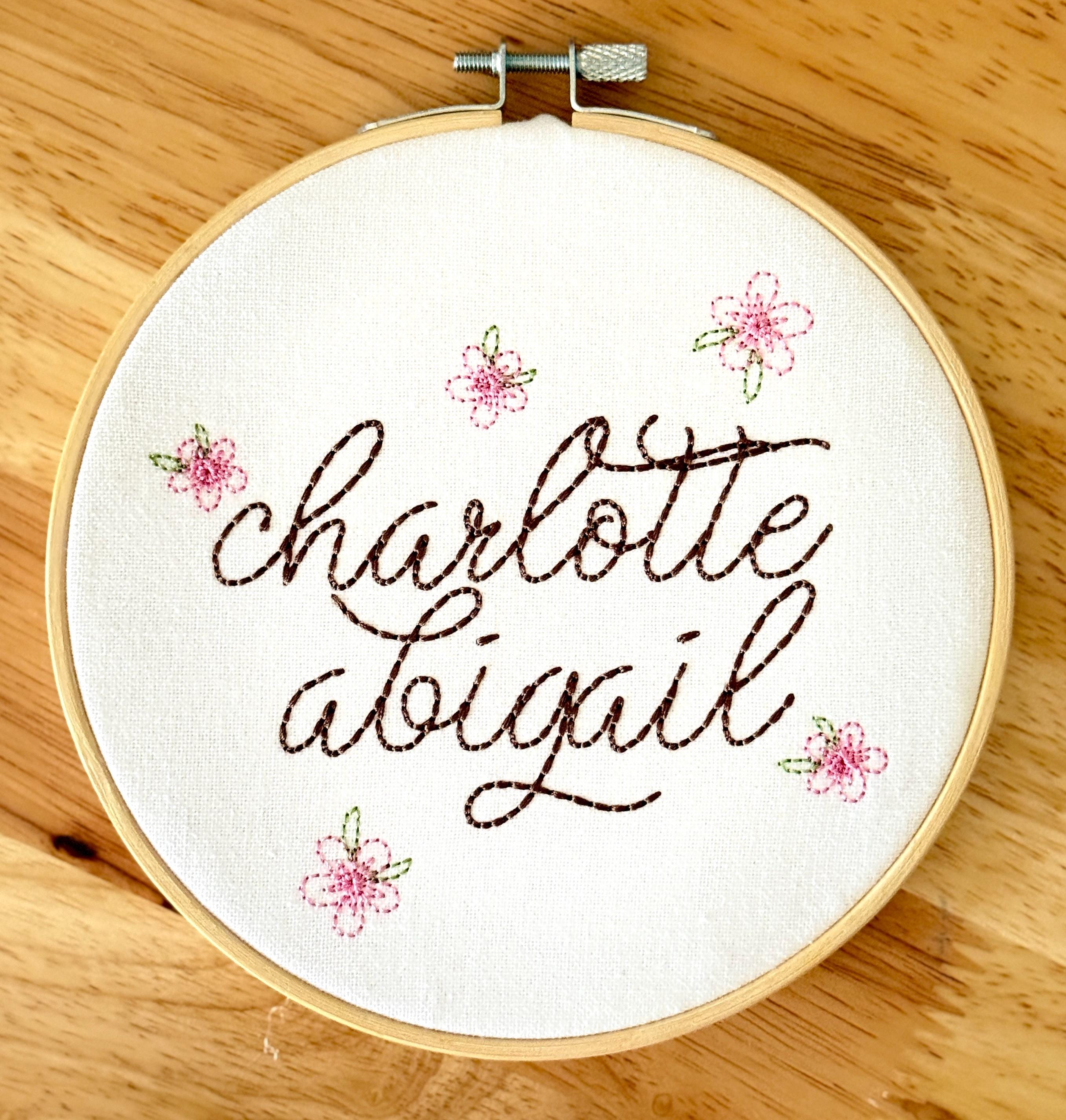 Light wood embroidery hoop art set on a wood table with the name Charlotte Abigail embroidered in dark brown. The hoop has five pink embroidered flowers around the name: Charlotte Abigail in cursive. It is stitched on white cotton fabric.