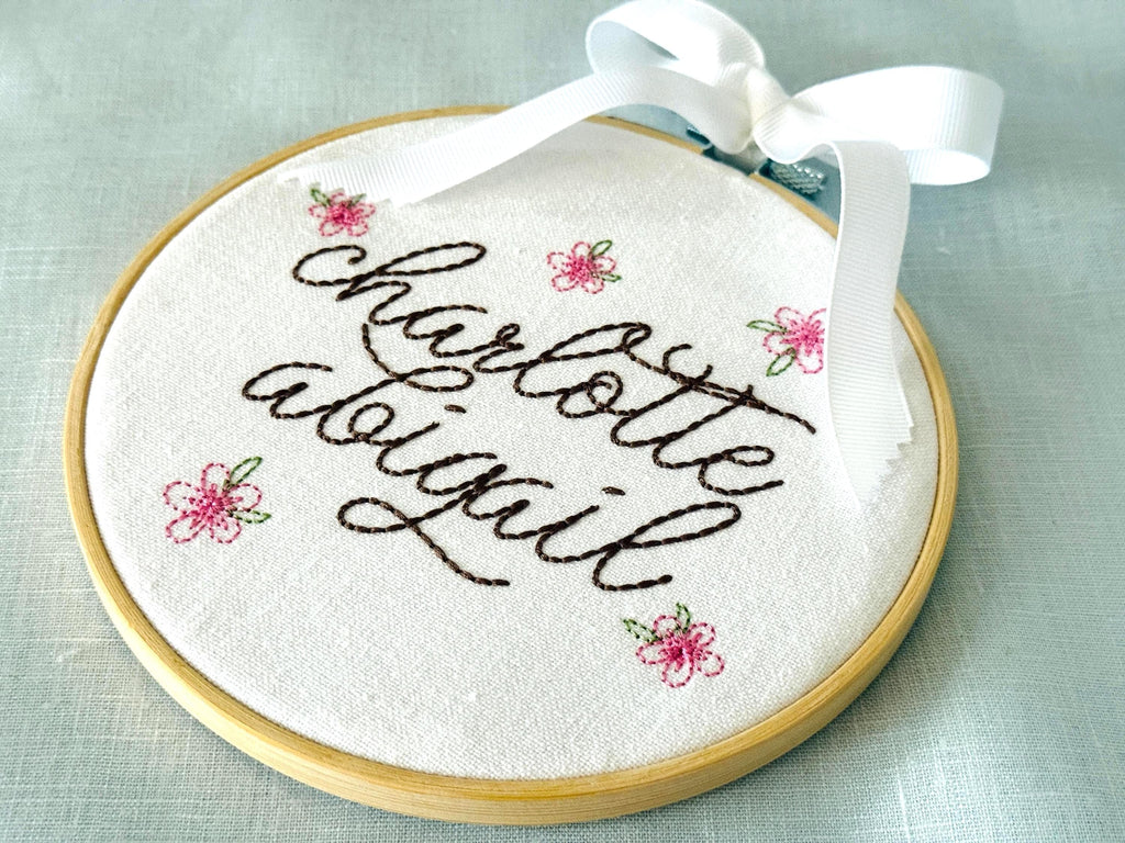 Light wood embroidery hoop art set on a light blue linen tablecloth with the name Charlotte Abigail embroidered in dark brown. The hoop has five pink embroidered flowers around the name: Charlotte Abigail in cursive. It is stitched on white cotton fabric.