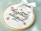Light wood embroidery hoop art set on a light blue linen tablecloth with the name Charlotte Abigail embroidered in dark brown. The hoop has five pink embroidered flowers around the name: Charlotte Abigail in cursive. It is stitched on white cotton fabric.