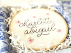A light wood embroidery hoop packaged in a white box with white crinkle paper and blue and white chinoiserie style tissue paper.  The hoop has five pink embroidered flowers around the name: Charlotte Abigail in cursive. It is stitched on white cotton fabric.