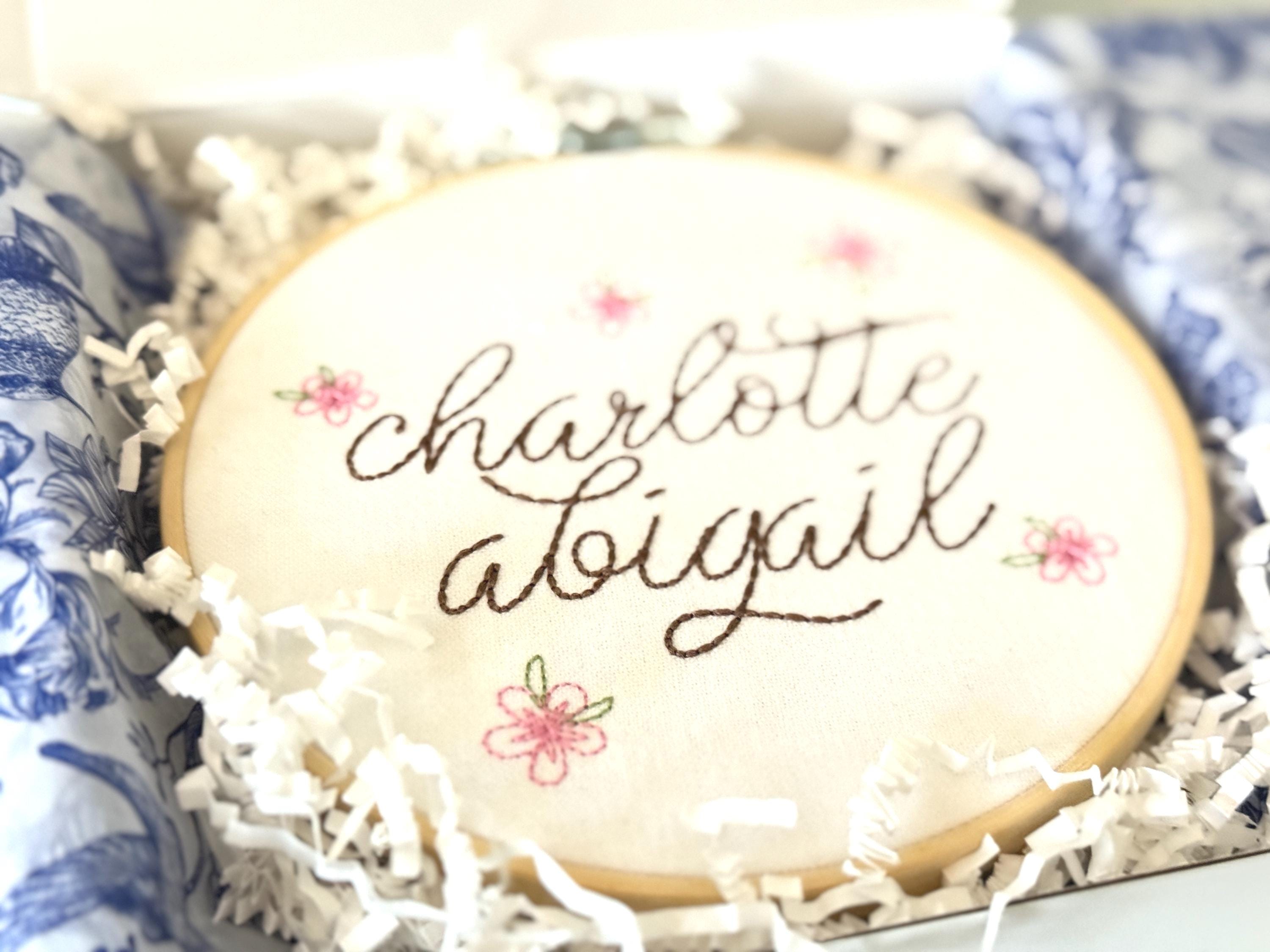 A light wood embroidery hoop packaged in a white box with white crinkle paper and blue and white chinoiserie style tissue paper.  The hoop has five pink embroidered flowers around the name: Charlotte Abigail in cursive. It is stitched on white cotton fabric.