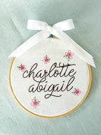 Light wood embroidery hoop art set on a light blue linen tablecloth with the name Charlotte Abigail embroidered in dark brown. The hoop has five pink embroidered flowers around the name: Charlotte Abigail in cursive. It is stitched on white cotton fabric.
