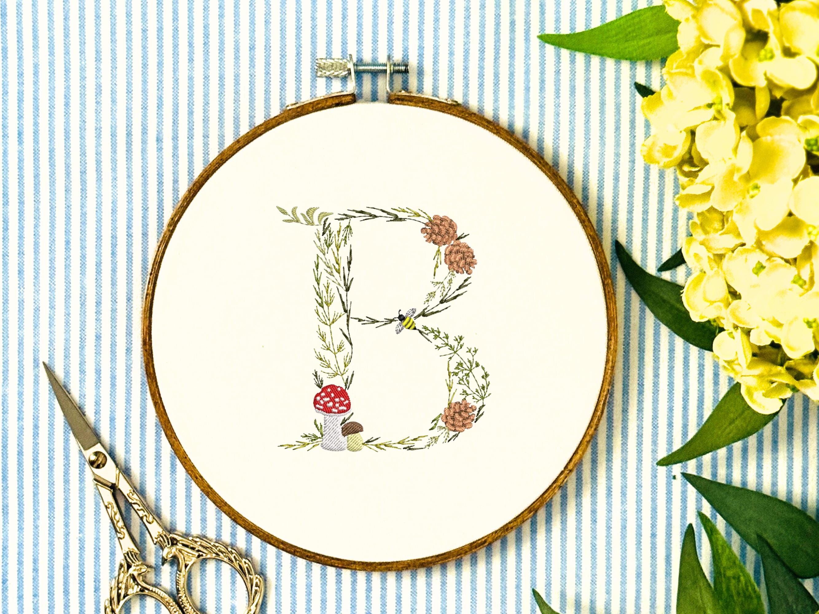 A letter B embroidered in a woodland theme with ferns, branches, acorn, a bee, and mushrooms in greens, browns, oranges, and yellow. This letter is stitched onto a white cotton background with a wooden embroidery hoop holding it. There are embroidery items like material, scissors, and thread around the photo. The photo is a top view.