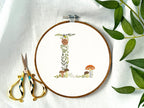 A letter L embroidered in a woodland theme with ferns, branches, acorn, a bee, and mushrooms in greens, browns, oranges, and yellow. This letter is stitched onto a white cotton background with a wooden embroidery hoop holding it. There are embroidery items like material, scissors, and thread around the photo. The photo is a top view.