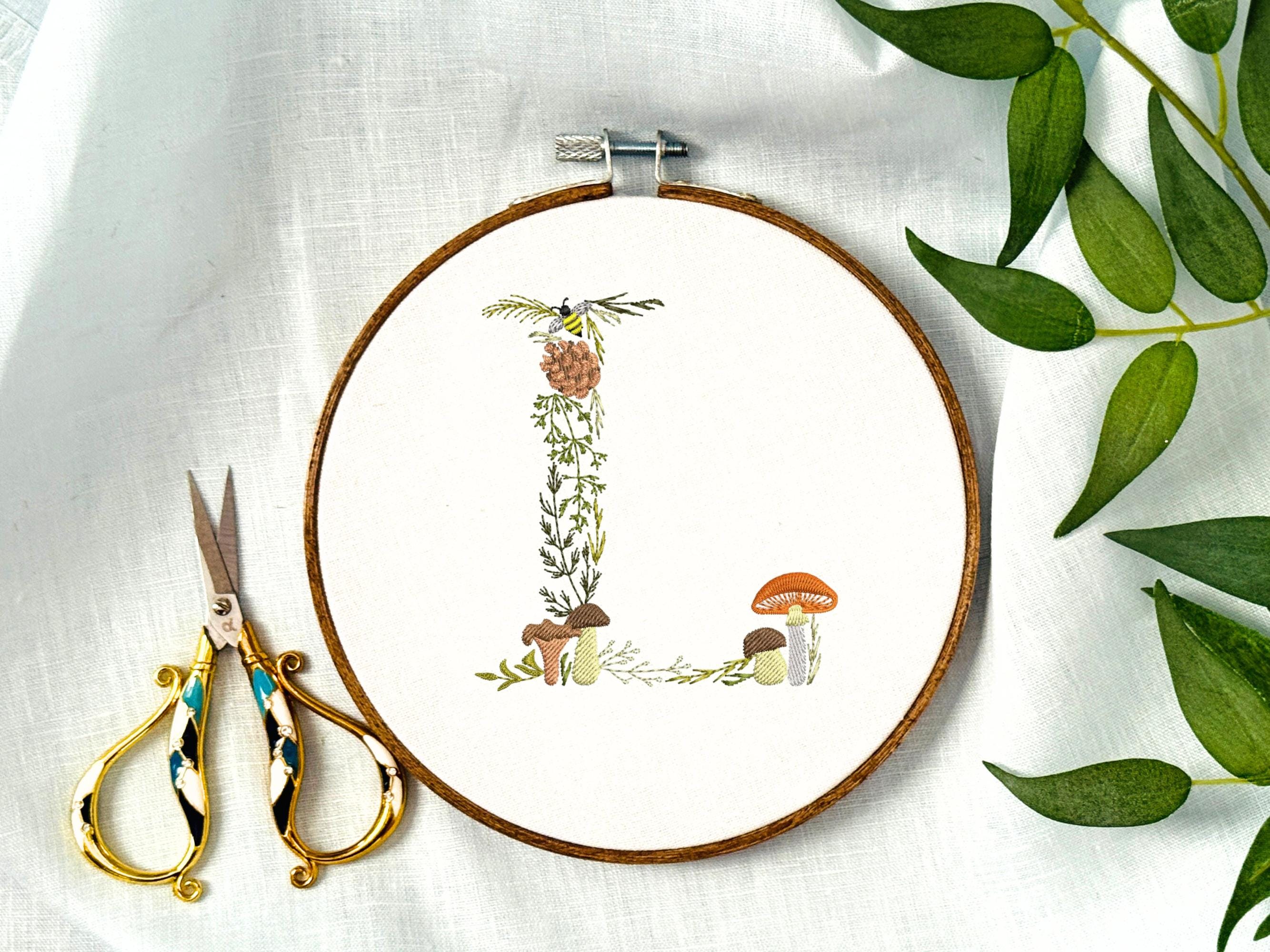 A letter L embroidered in a woodland theme with ferns, branches, acorn, a bee, and mushrooms in greens, browns, oranges, and yellow. This letter is stitched onto a white cotton background with a wooden embroidery hoop holding it. There are embroidery items like material, scissors, and thread around the photo. The photo is a top view.