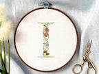 A letter I embroidered in a woodland theme with ferns, branches, acorn, a bee, and mushrooms in greens, browns, oranges, and yellow. This letter is stitched onto a white cotton background with a wooden embroidery hoop holding it. There are embroidery items like material, scissors, and thread around the photo. The photo is a top view.