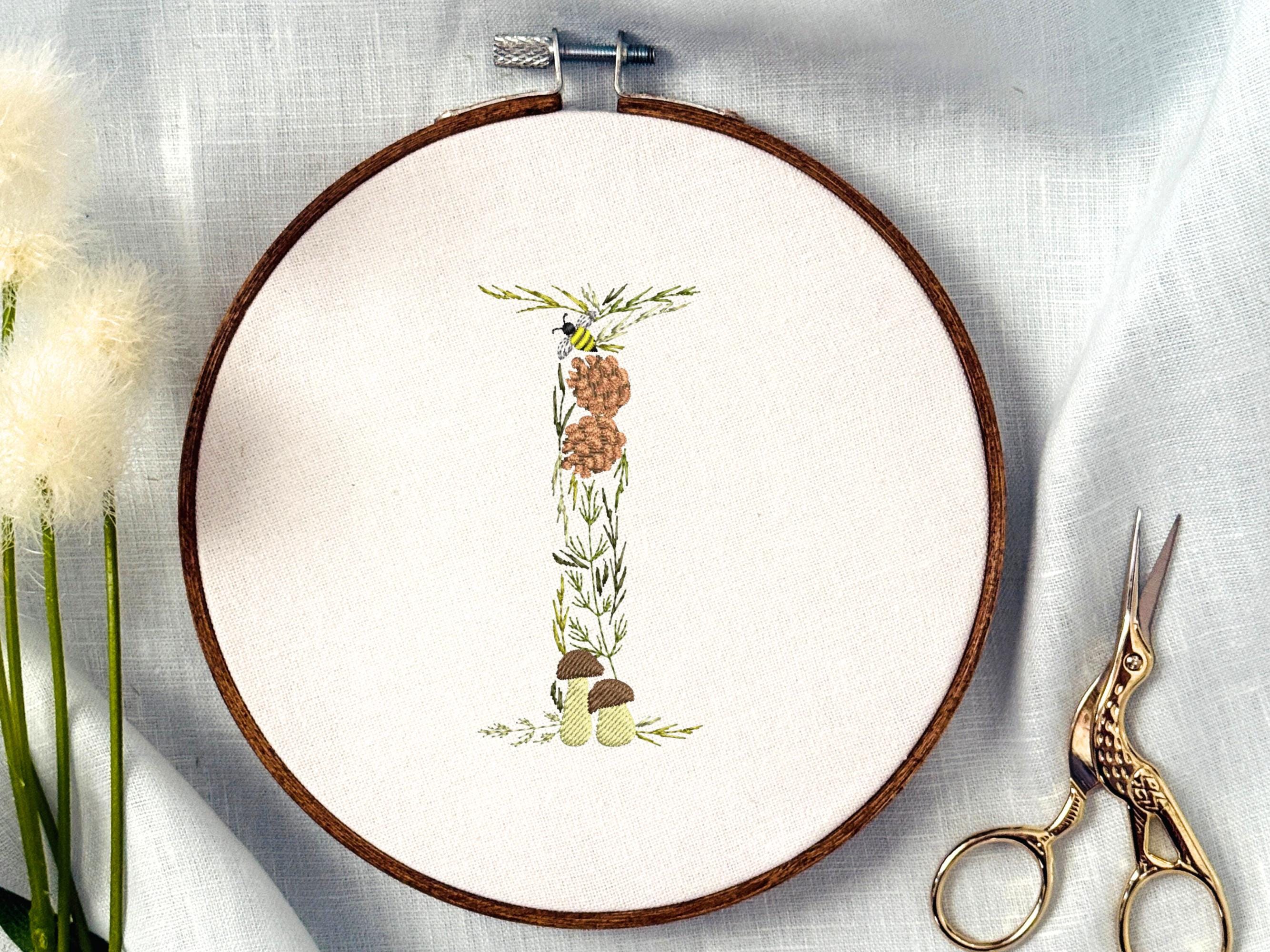 A letter I embroidered in a woodland theme with ferns, branches, acorn, a bee, and mushrooms in greens, browns, oranges, and yellow. This letter is stitched onto a white cotton background with a wooden embroidery hoop holding it. There are embroidery items like material, scissors, and thread around the photo. The photo is a top view.