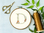 A letter D embroidered in a woodland theme with ferns, branches, acorn, a bee, and mushrooms in greens, browns, oranges, and yellow. This letter is stitched onto a white cotton background with a wooden embroidery hoop holding it. There are embroidery items like material, scissors, and thread around the photo. The photo is a top view.