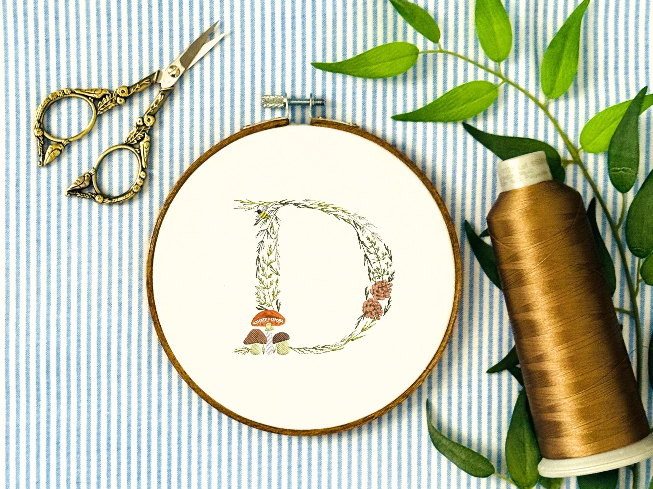 A letter D embroidered in a woodland theme with ferns, branches, acorn, a bee, and mushrooms in greens, browns, oranges, and yellow. This letter is stitched onto a white cotton background with a wooden embroidery hoop holding it. There are embroidery items like material, scissors, and thread around the photo. The photo is a top view.