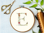 A letter E embroidered in a woodland theme with ferns, branches, acorn, a bee, and mushrooms in greens, browns, oranges, and yellow. This letter is stitched onto a white cotton background with a wooden embroidery hoop holding it. There are embroidery items like material, scissors, and thread around the photo. The photo is a top view.