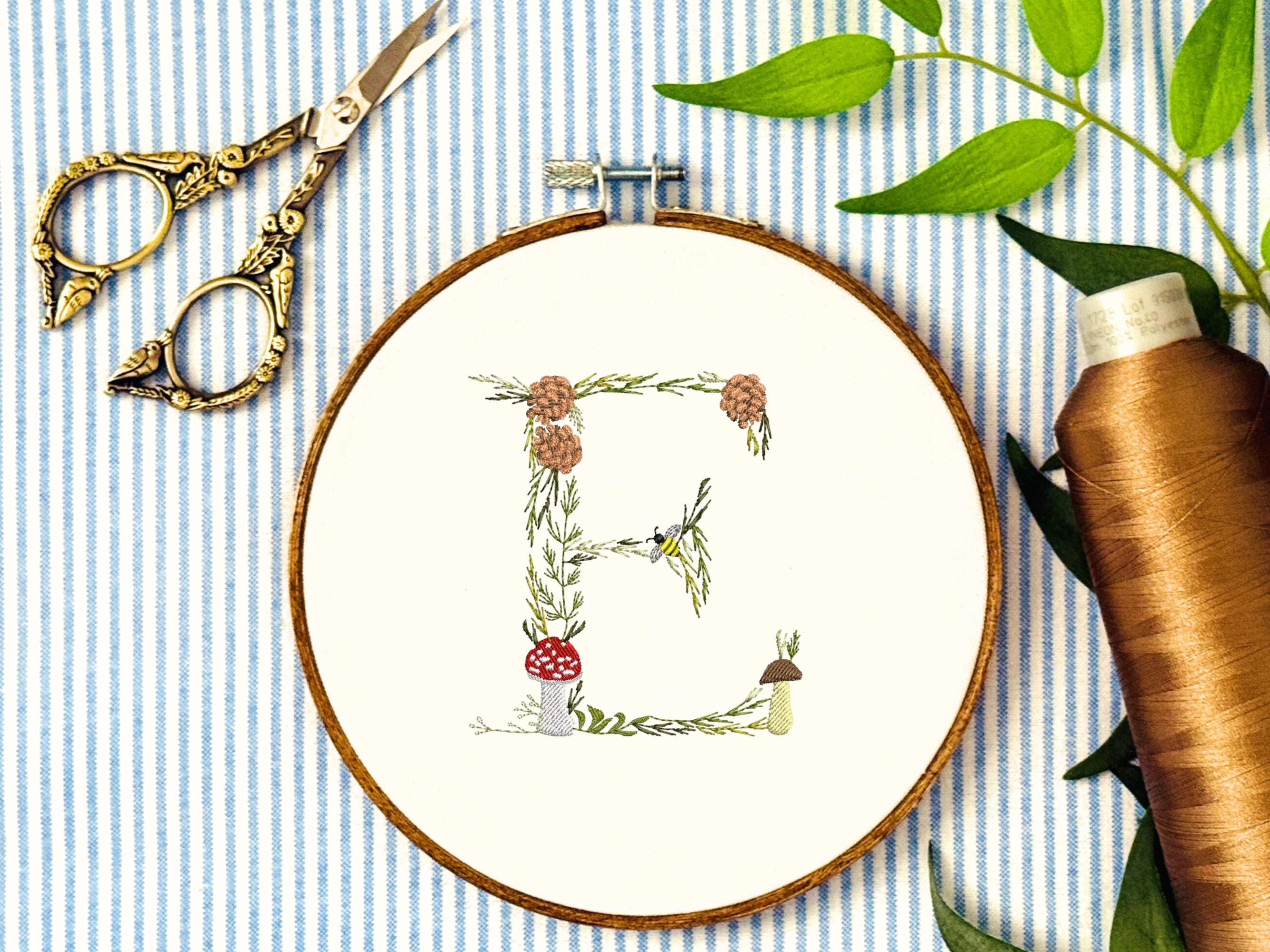 A letter E embroidered in a woodland theme with ferns, branches, acorn, a bee, and mushrooms in greens, browns, oranges, and yellow. This letter is stitched onto a white cotton background with a wooden embroidery hoop holding it. There are embroidery items like material, scissors, and thread around the photo. The photo is a top view.