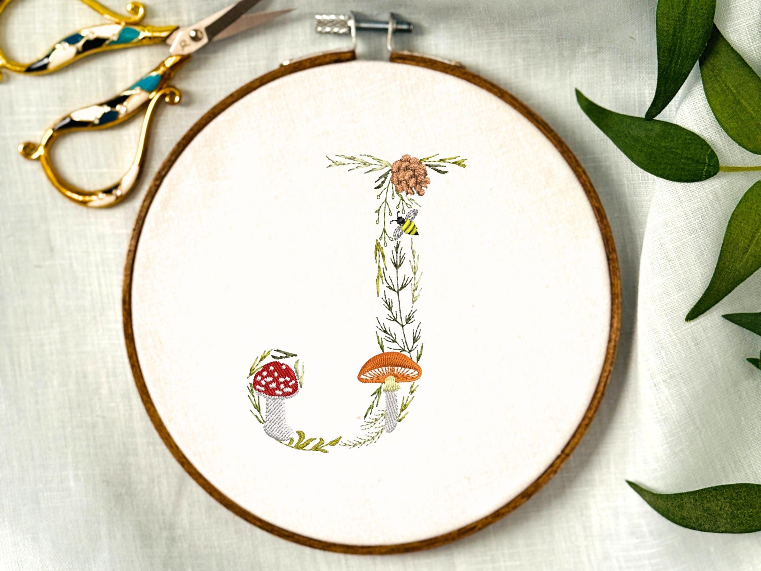 A letter J embroidered in a woodland theme with ferns, branches, acorn, a bee, and mushrooms in greens, browns, oranges, and yellow. This letter is stitched onto a white cotton background with a wooden embroidery hoop holding it. There are embroidery items like material, scissors, and thread around the photo. The photo is a top view.