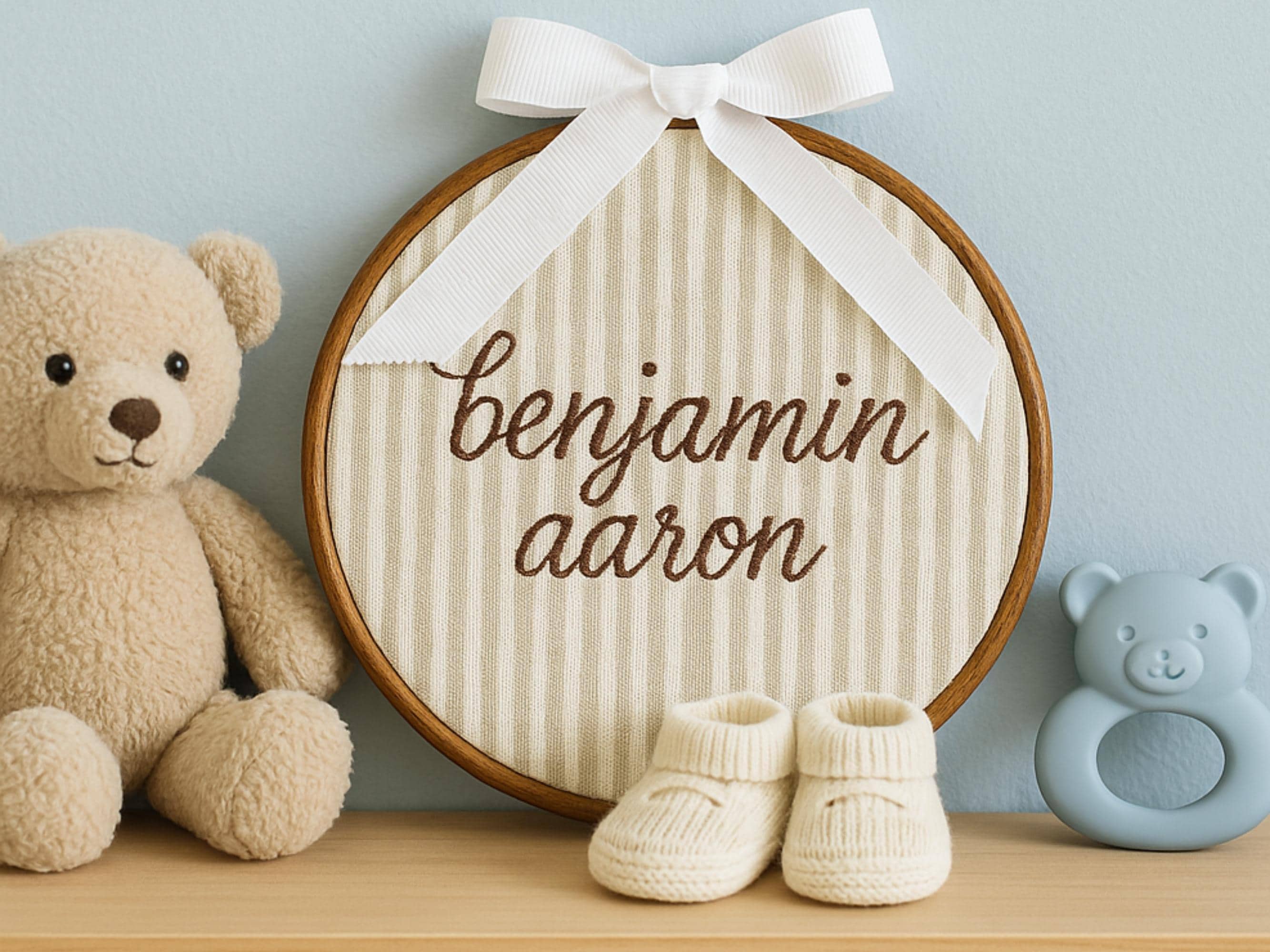 A walnut color hoop with tan and white pin stripes with the name Benjamin Aaron stitched in brown thread. The hoop is on a shelf next to a light brown teddy bear, baby shoes, and a blue rattle. The hoop is leaning on the wall behind with a white ribbon on top.