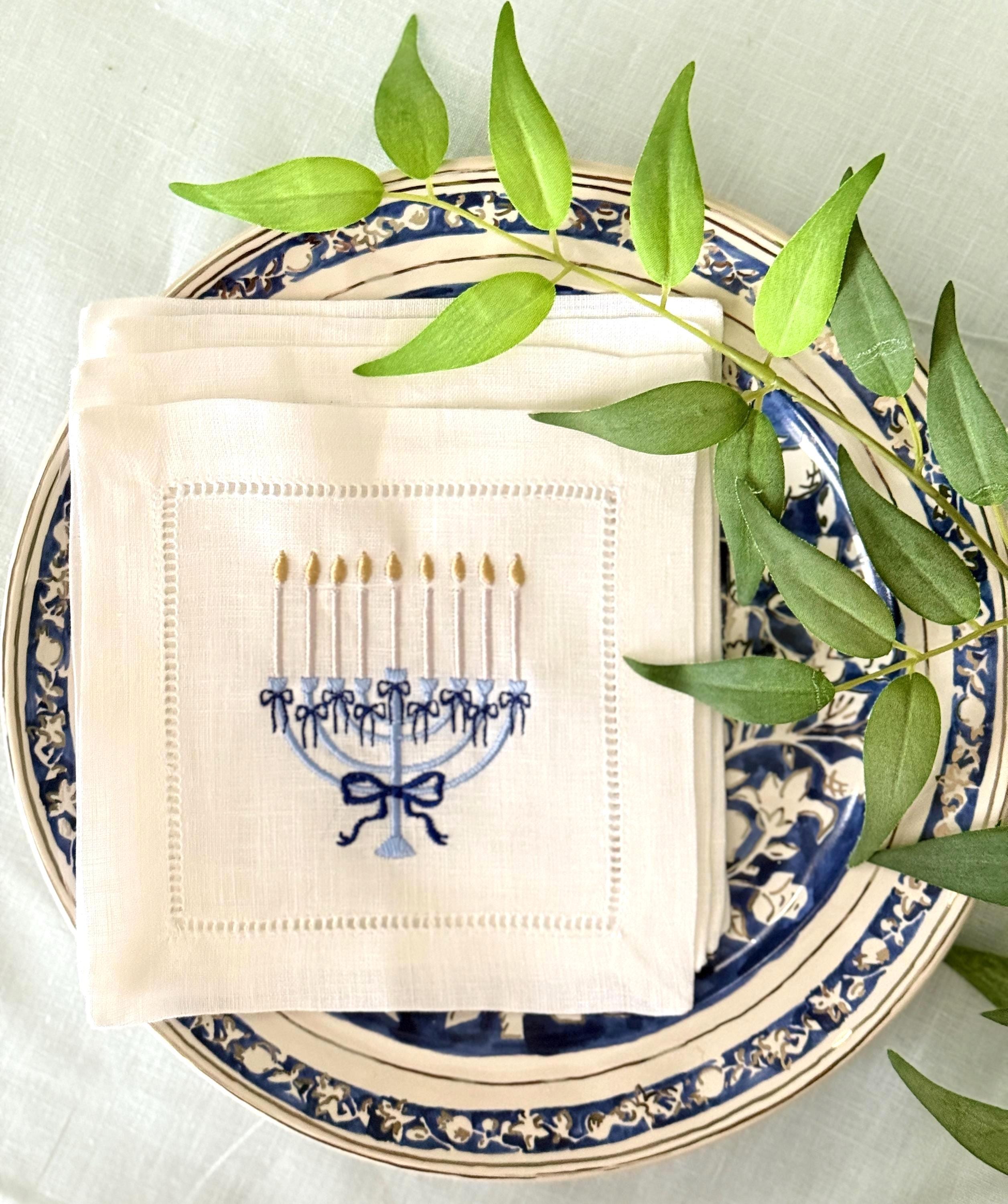 A 6x6 inch 100% linen cocktail napkin, set of 4 embroidered with a light blue Hanukkah Menorah that has dark blue ribbons and gold thread flames set on a Hanukkah plate set on a light blue linen tablecloth with a green branch with leaves.