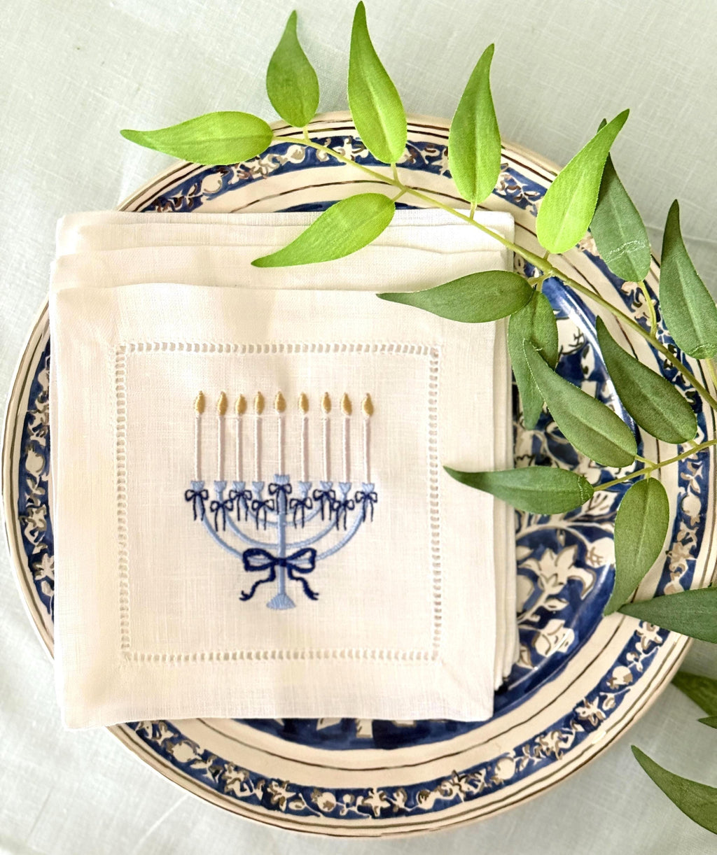A 6x6 inch 100% linen cocktail napkin, set of 4 embroidered with a light blue Hanukkah Menorah that has dark blue ribbons and gold thread flames set on a Hanukkah plate set on a light blue linen tablecloth with a green branch with leaves.