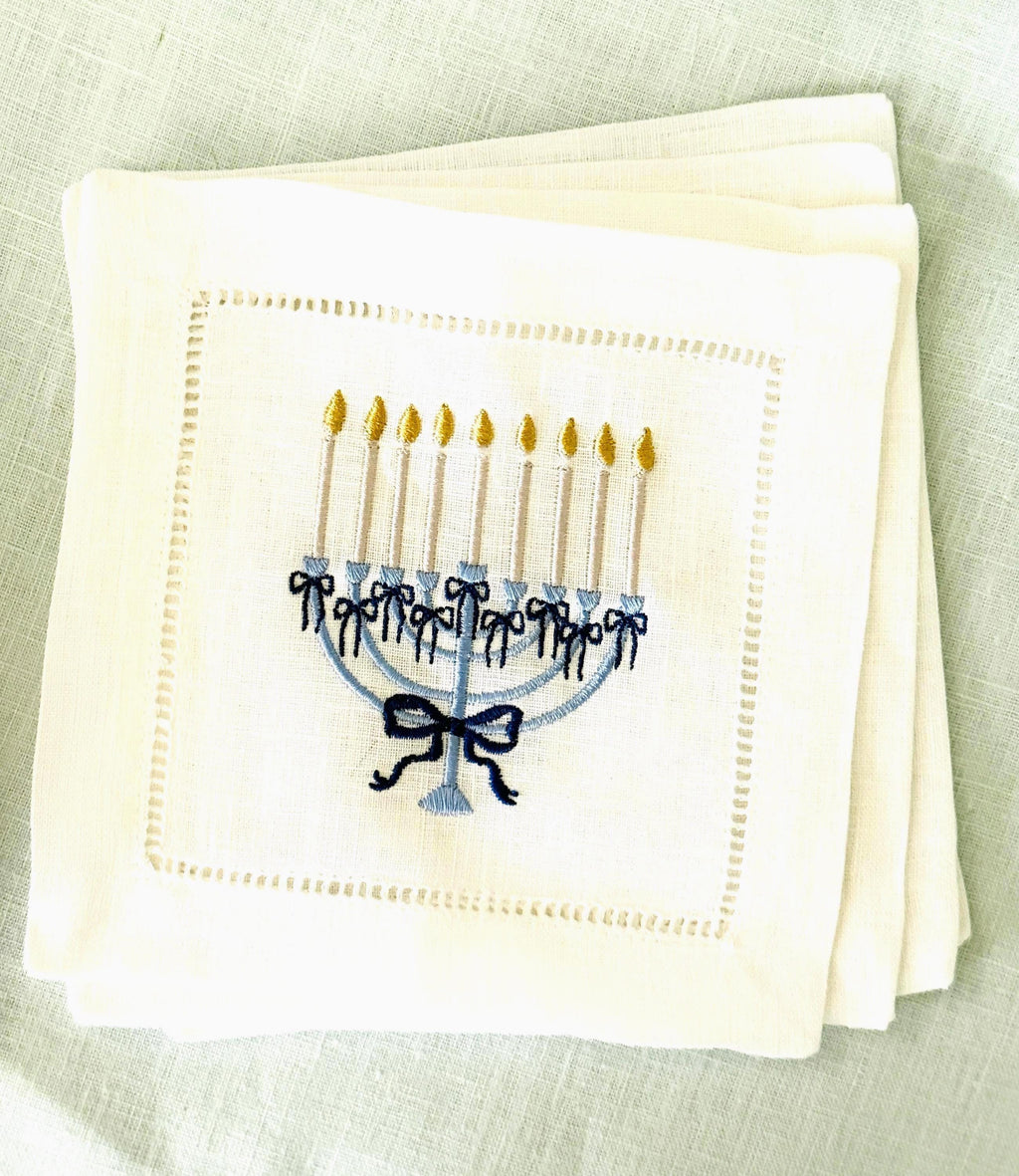 A 6x6 inch 100% linen cocktail napkin, set of 4 embroidered with a light blue Hanukkah Menorah that has dark blue ribbons and gold thread flames set on a Hanukkah plate set on a light blue linen tablecloth.