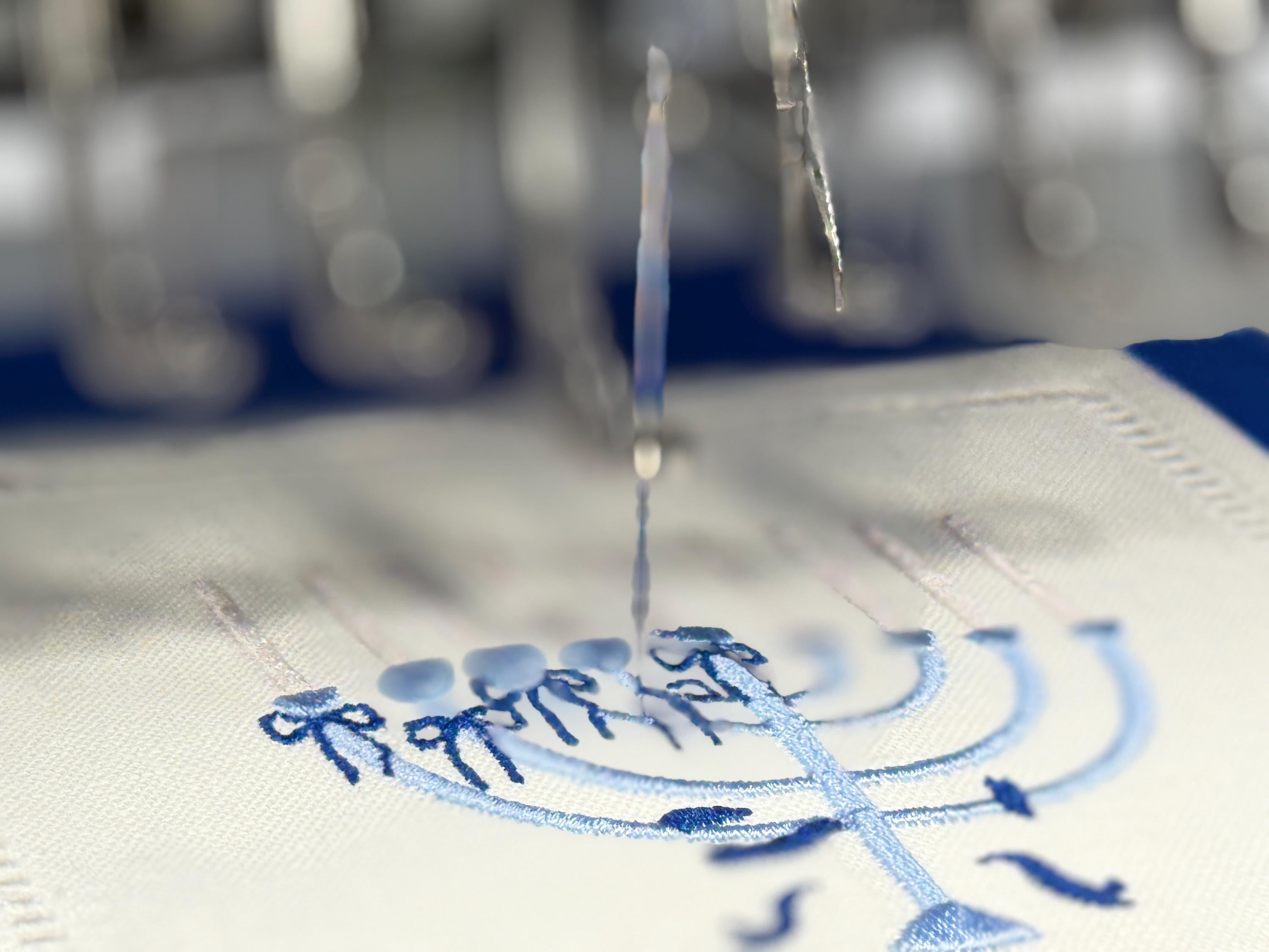 An action shot of A 6x6 inch 100% linen cocktail napkin being embroidered with a light blue Hanukkah Menorah that has dark blue ribbons and gold thread flames.