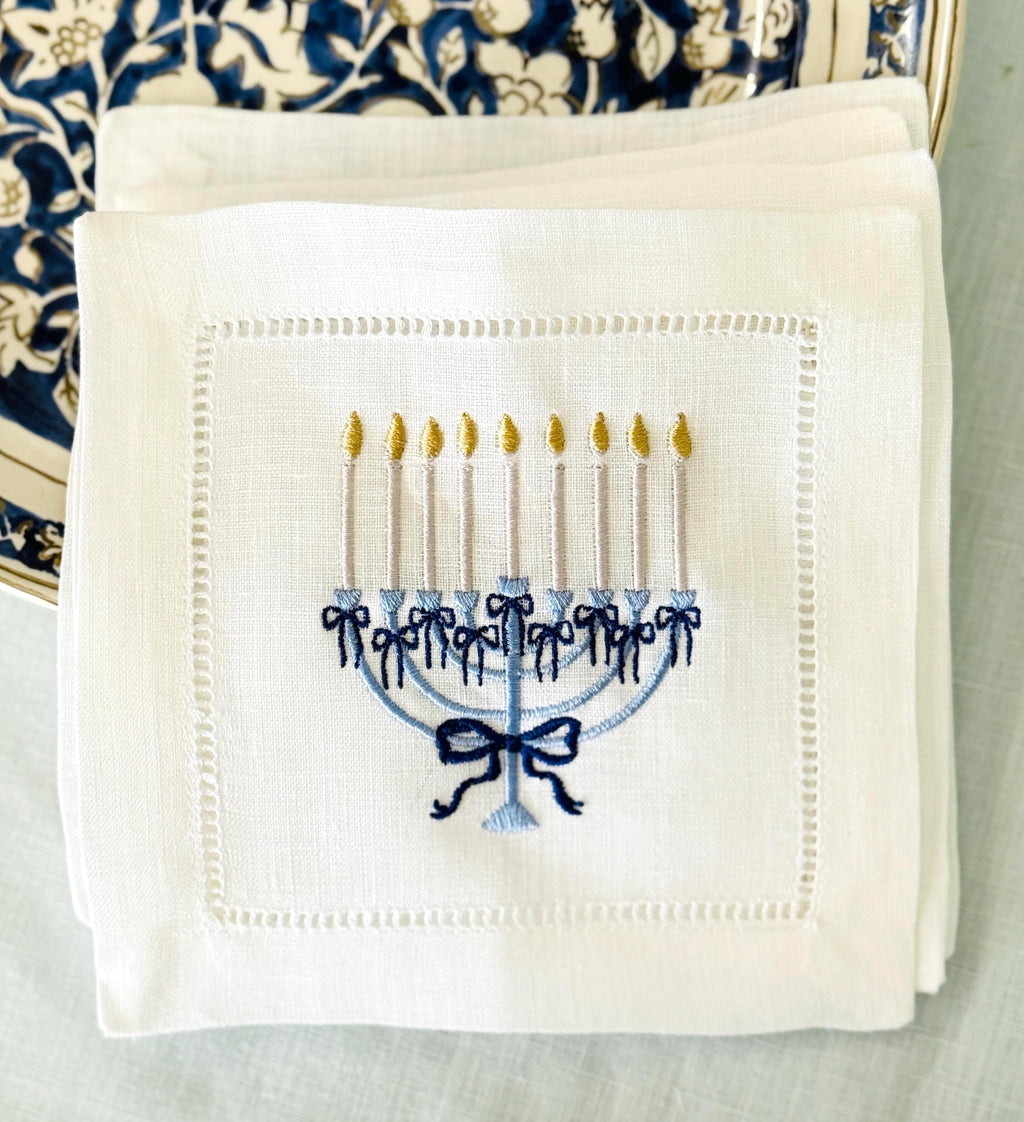 A 6x6 inch 100% linen cocktail napkin, set of 4 embroidered with a light blue Hanukkah Menorah that has dark blue ribbons and gold thread flames set on a Hanukkah plate set on a light blue linen tablecloth.
