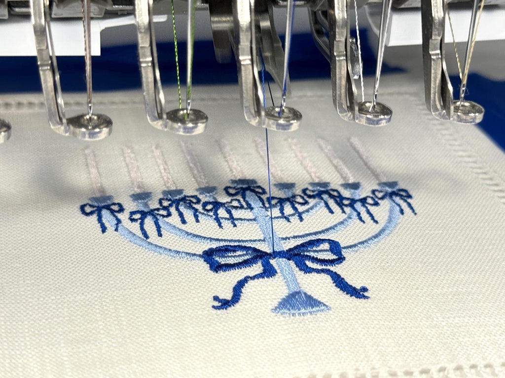 A 6x6 inch 100% linen cocktail napkin, set of 4 embroidered with a light blue Hanukkah Menorah that has dark blue ribbons and gold thread flames in the process of being embroidered.