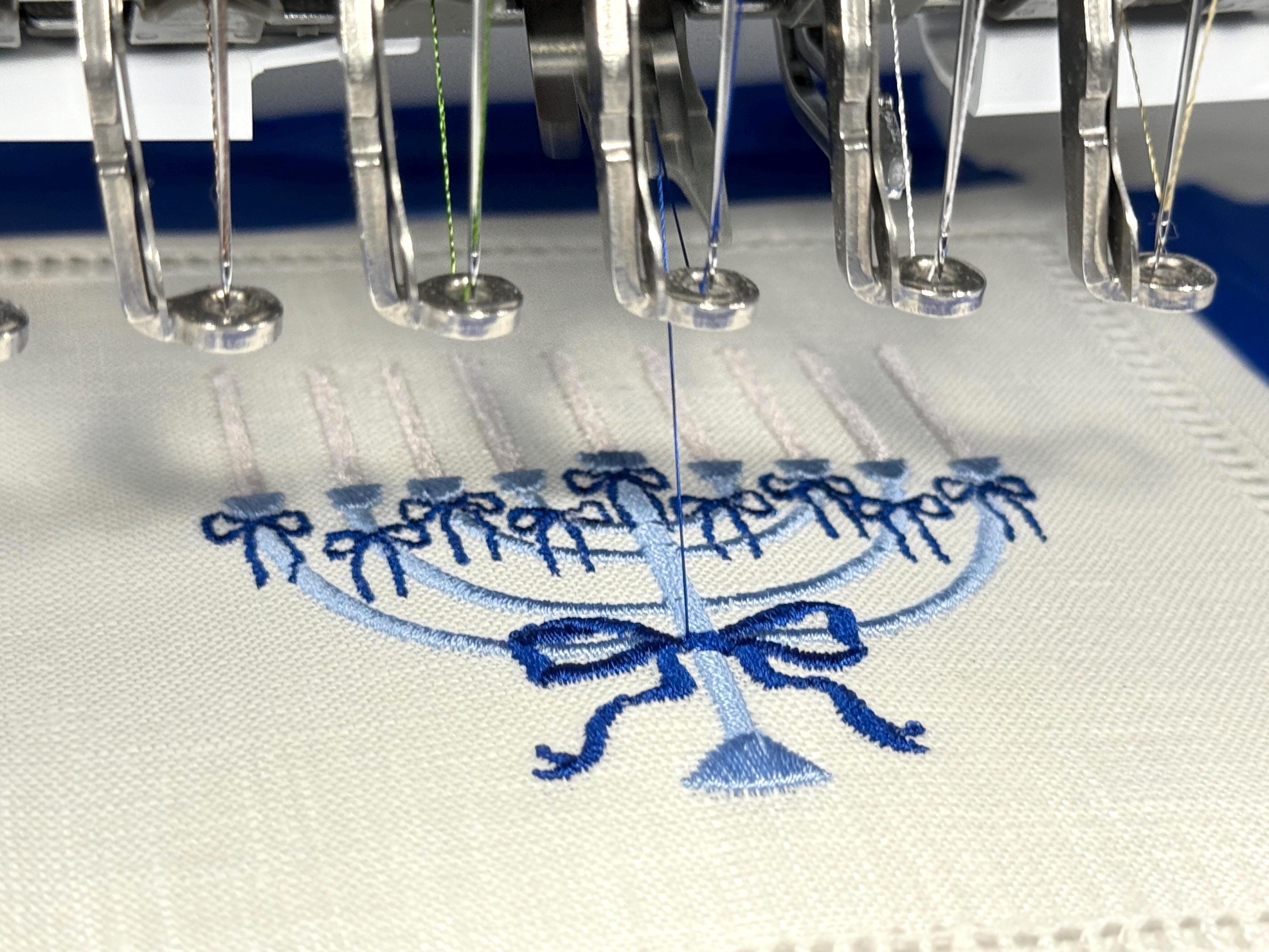 A 6x6 inch 100% linen cocktail napkin, set of 4 embroidered with a light blue Hanukkah Menorah that has dark blue ribbons and gold thread flames in the process of being embroidered.