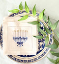 A 6x6 inch 100% linen cocktail napkin, set of 4 embroidered with a light blue Hanukkah Menorah that has dark blue ribbons and gold thread flames set on a Hanukkah plate with a green branch.