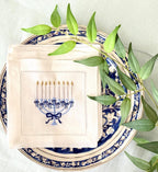 A 6x6 inch 100% linen cocktail napkin, set of 4 embroidered with a light blue Hanukkah Menorah that has dark blue ribbons and gold thread flames set on a Hanukkah plate with a green branch.