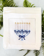 A 6x6 inch 100% linen cocktail napkin, set of 4 embroidered with a light blue Hanukkah Menorah that has dark blue ribbons and gold thread flames set on a Hanukkah plate set on a light blue linen tablecloth with a green fern branch under the napkins.